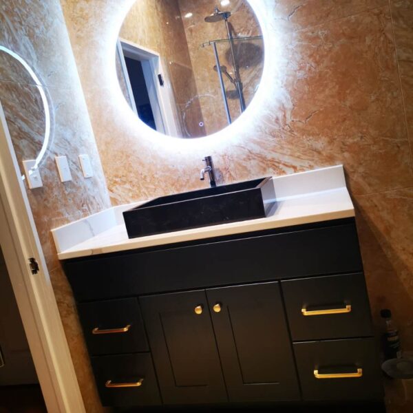 Modern bathroom vanity with illuminated round mirror and marble walls.