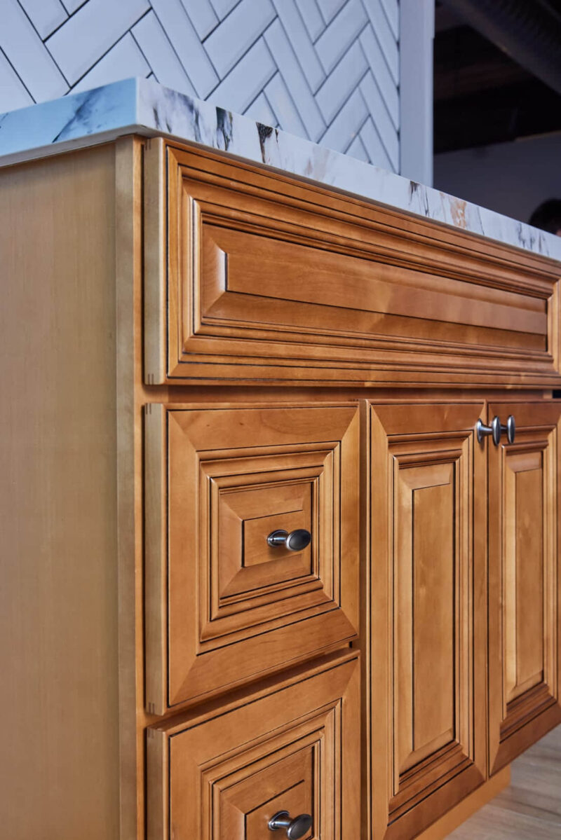 Wooden kitchen cabinet with marble countertop and herringbone tile backsplash.