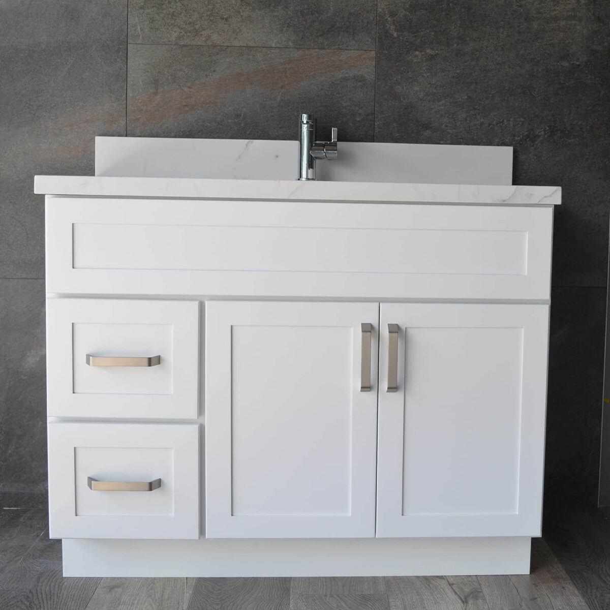 White bathroom vanity with drawers and sink against dark tiled wall.
