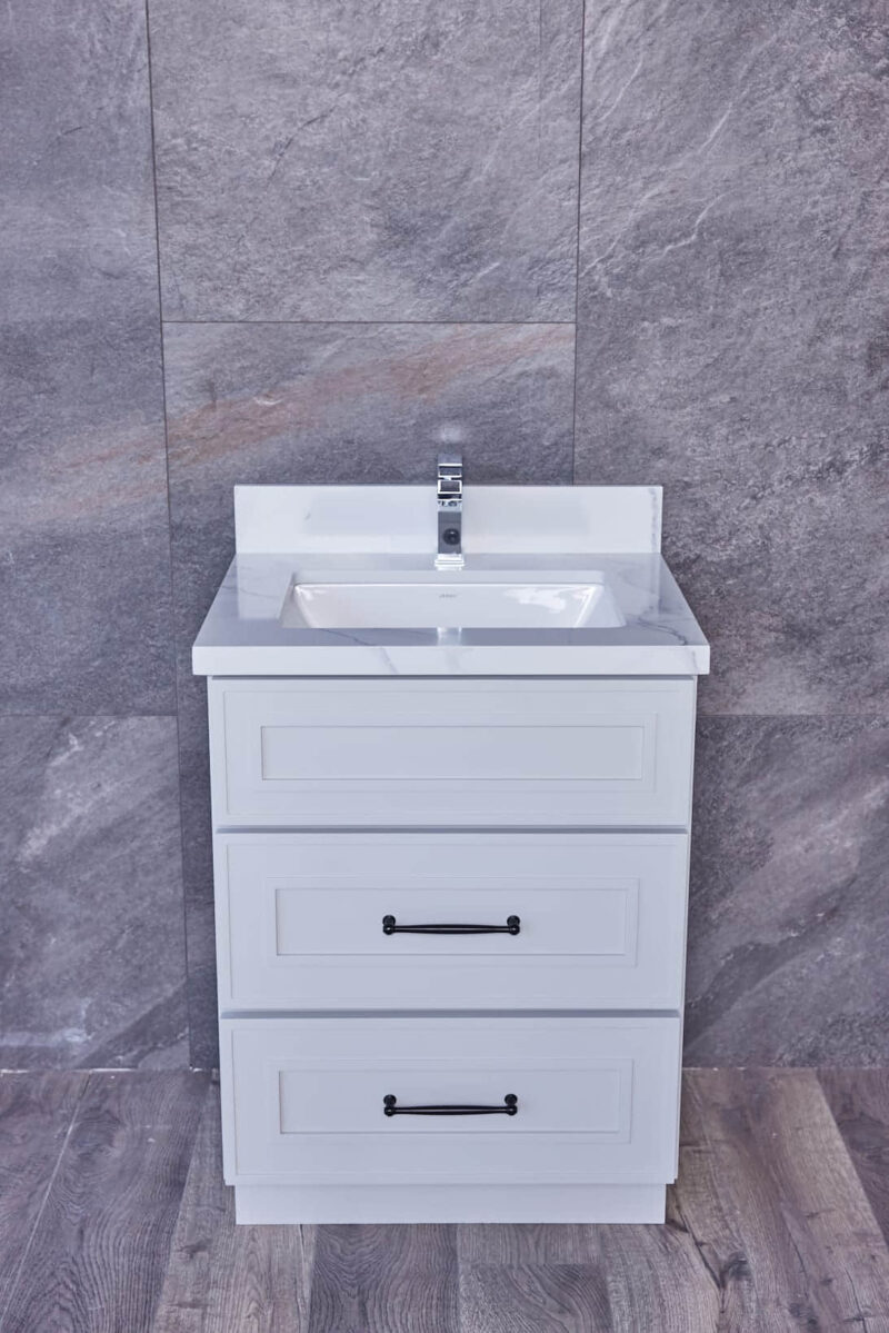 White bathroom vanity with three drawers and a sink, against a gray tiled wall.