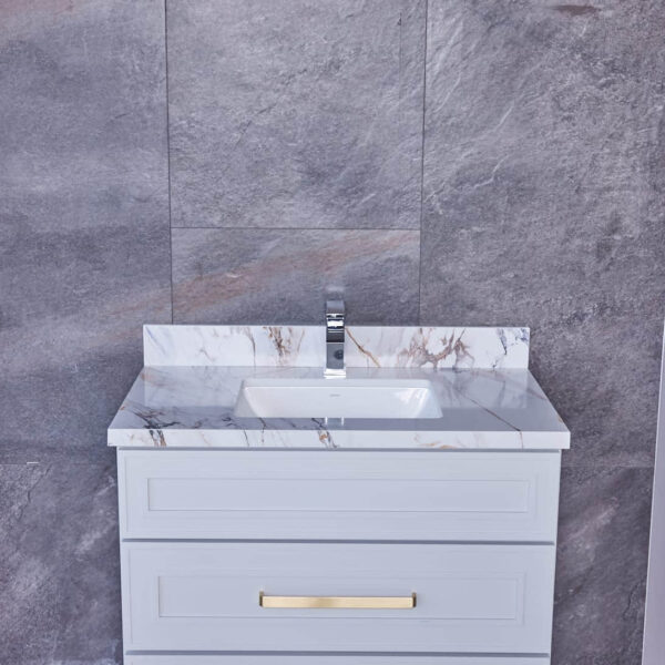 White bathroom vanity with marble countertop and gold handles against gray tile wall.