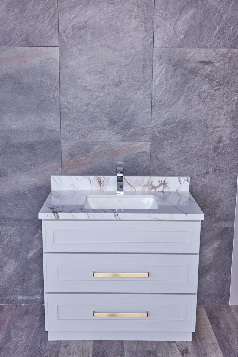 White bathroom vanity with marble countertop and gold handles against gray tile wall.