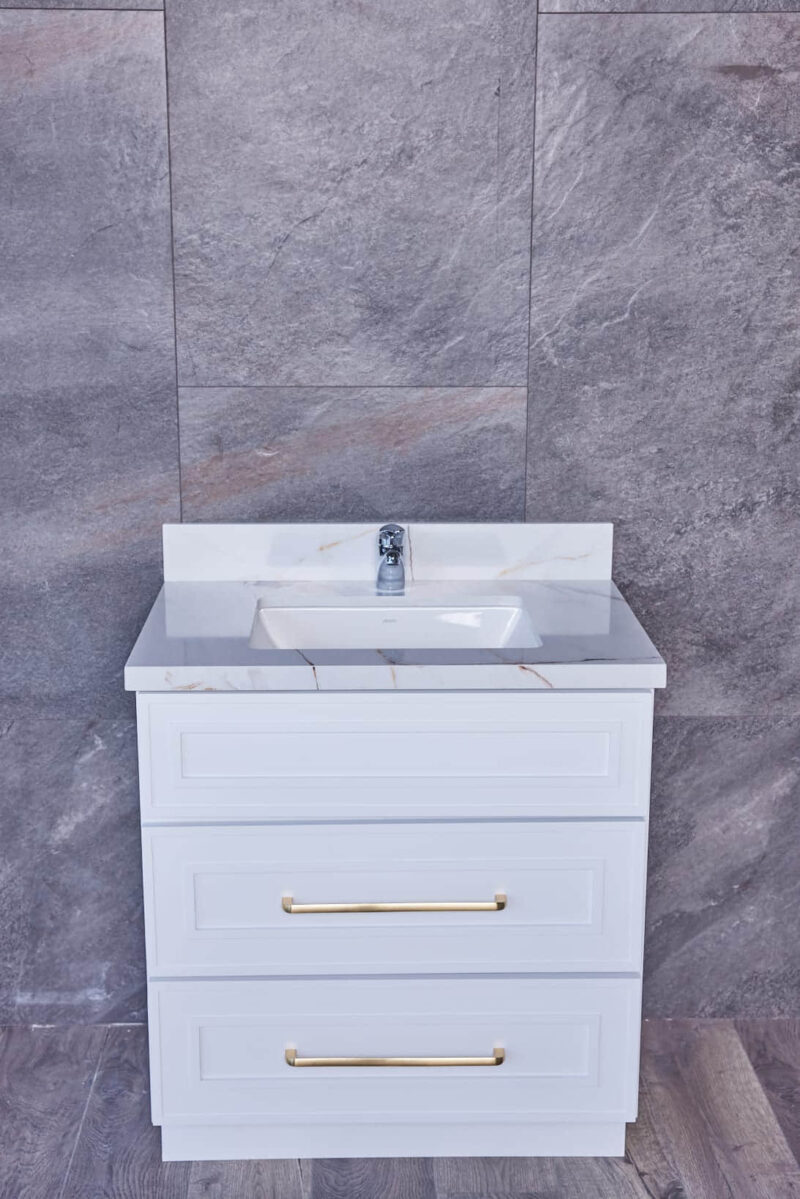 White bathroom vanity with marble countertop and gold handles against gray tile wall.