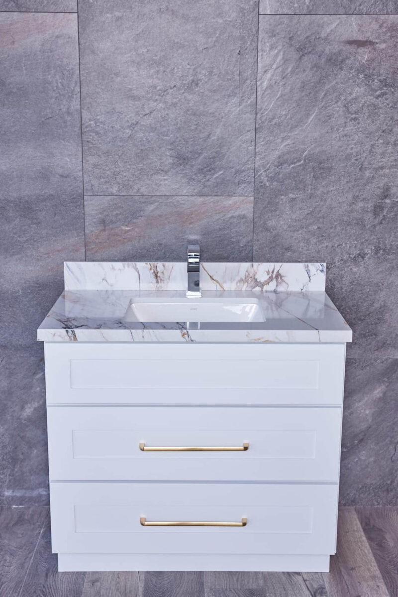 White bathroom vanity with marble countertop and gold handles against gray tiled wall.
