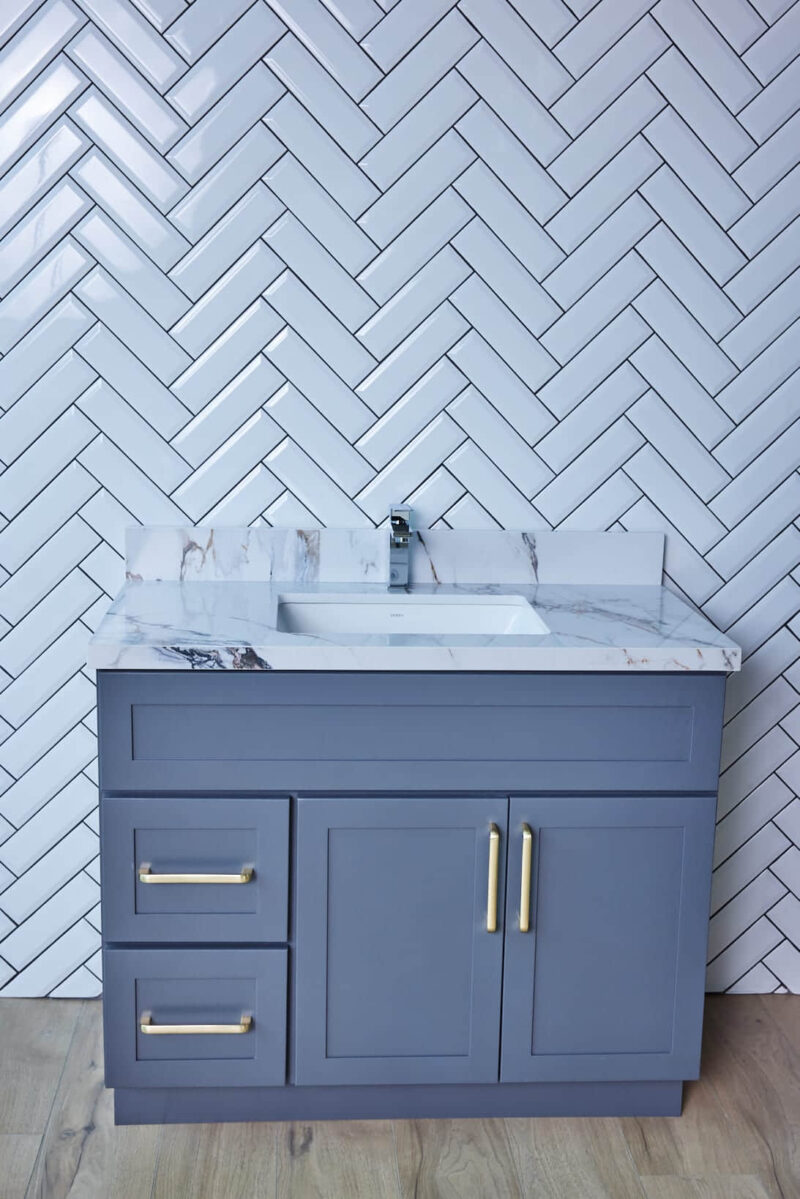 Gray bathroom vanity with marble countertop and herringbone tile backsplash.