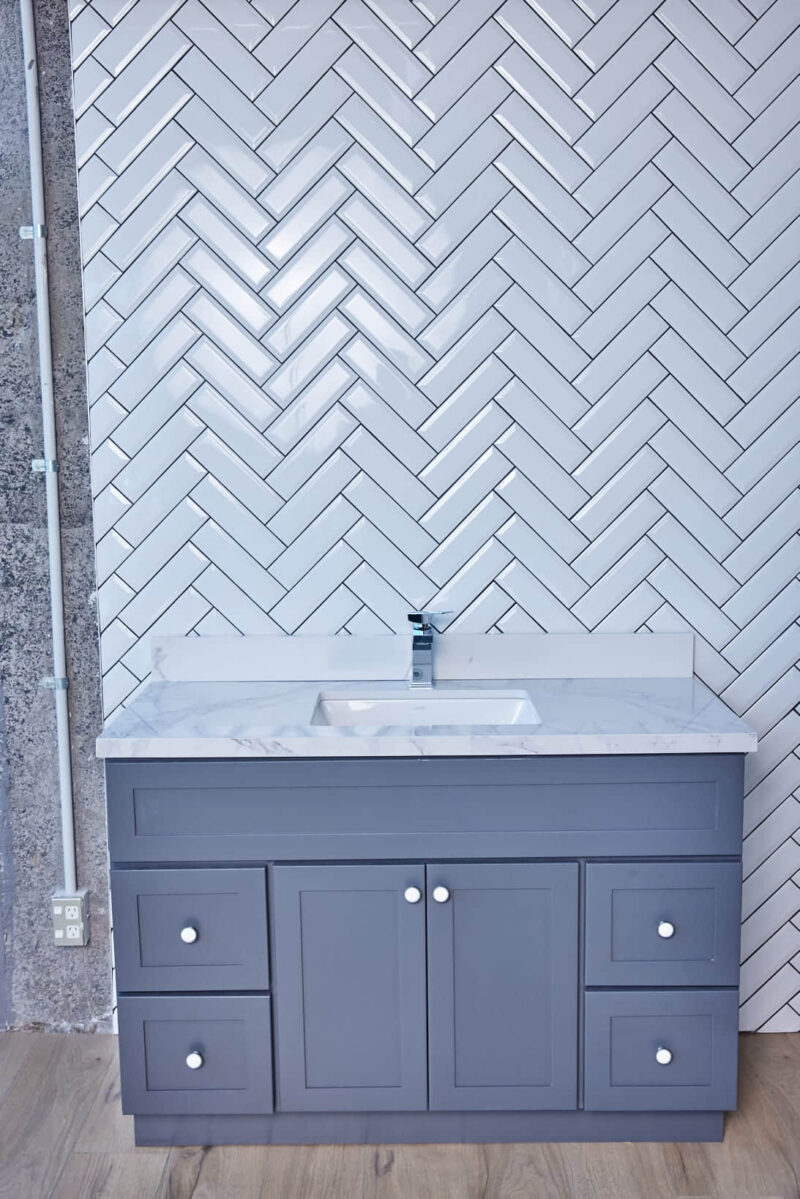 Gray bathroom vanity with marble countertop and herringbone tile backsplash.