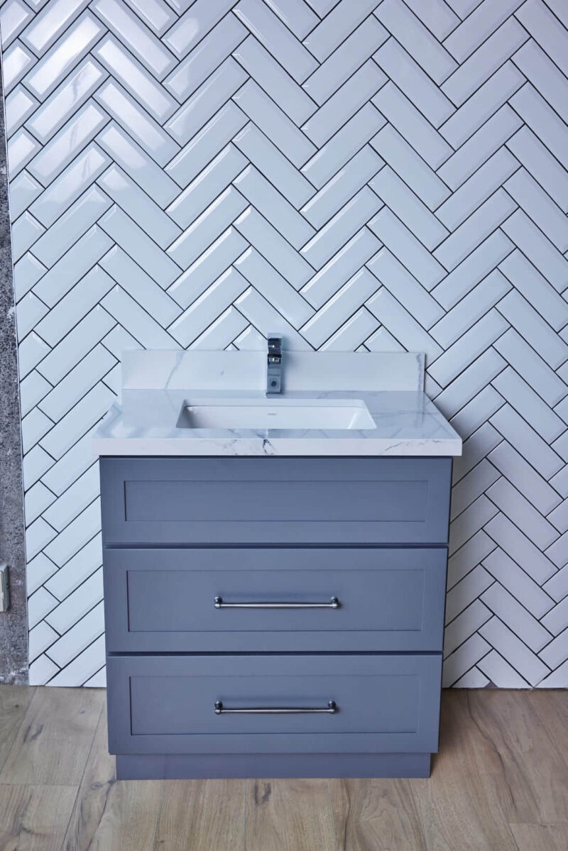 Gray bathroom vanity with white sink against herringbone tile wall.