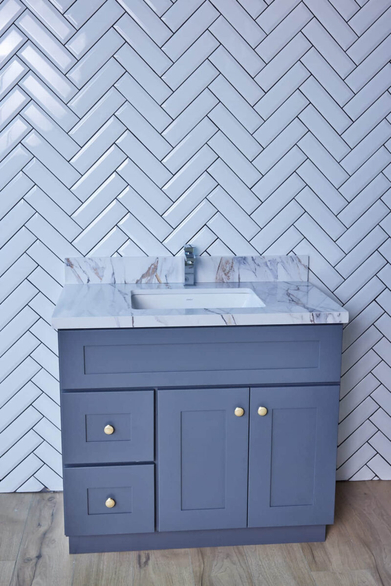 Gray bathroom vanity with marble countertop against herringbone tile wall.