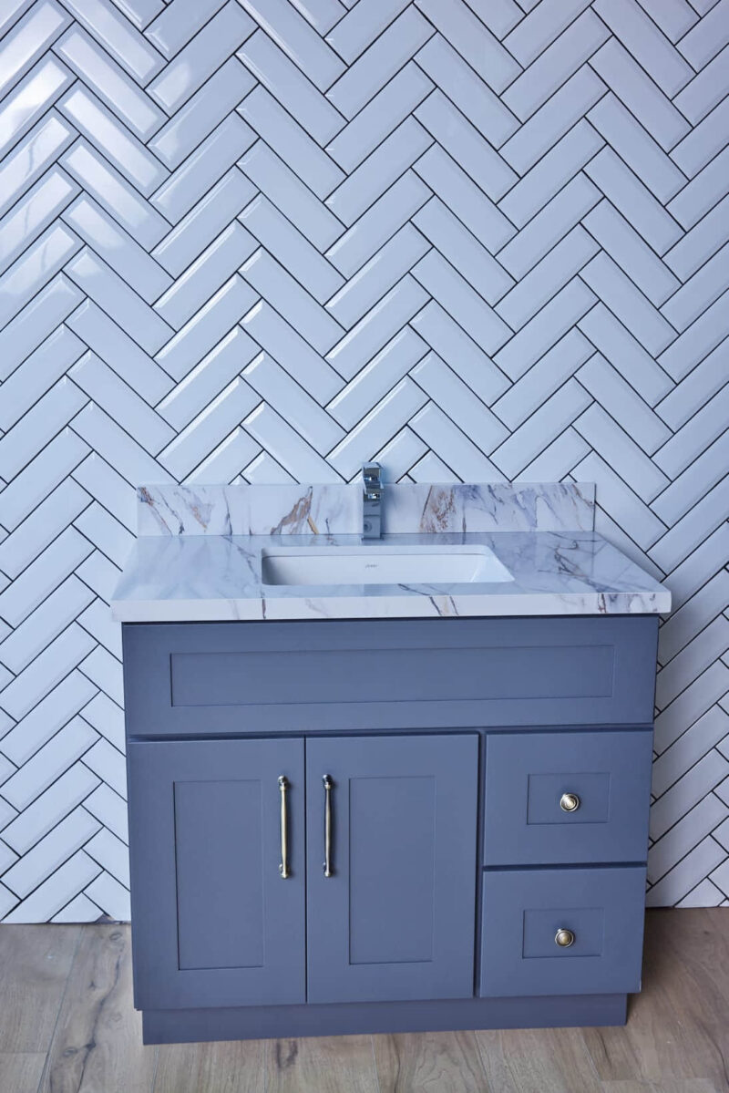 Gray bathroom vanity with marble countertop and herringbone tile backsplash.