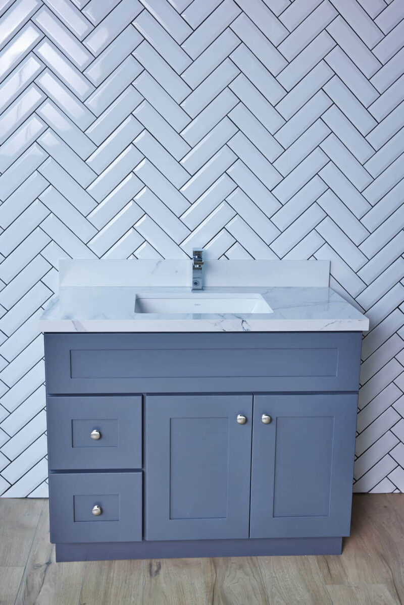 Gray bathroom vanity with marble countertop against herringbone tile wall.