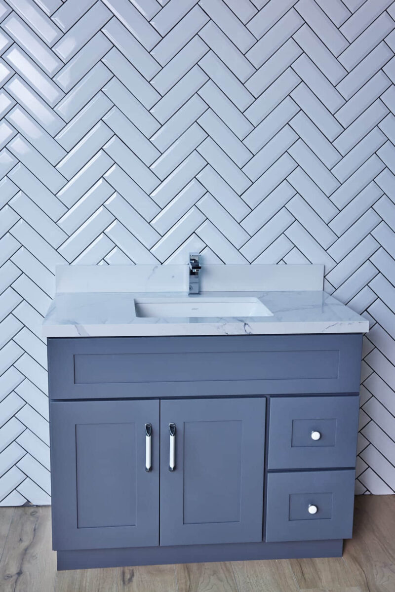 Gray bathroom vanity with marble countertop and herringbone tile backsplash.