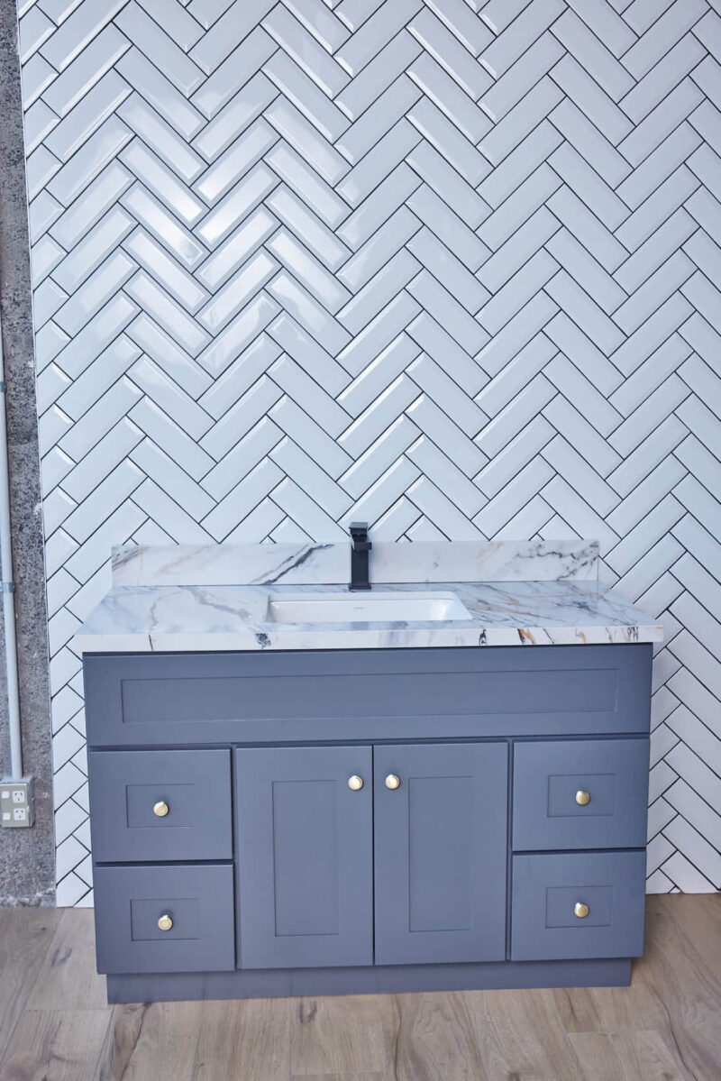 Gray bathroom vanity with marble countertop and herringbone tile backsplash.