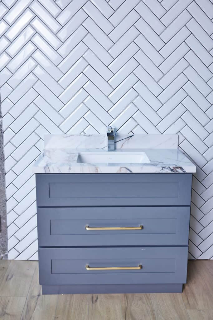 Gray bathroom vanity with marble countertop and herringbone tile backsplash.