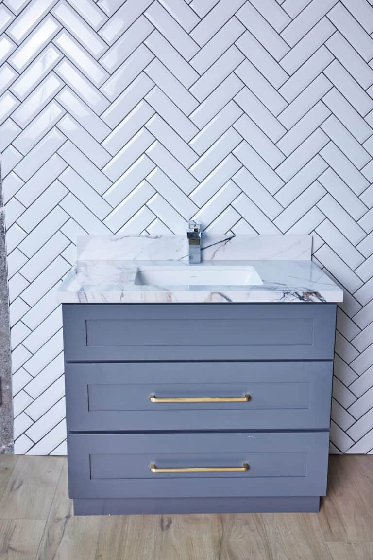 Gray bathroom vanity with marble countertop and herringbone tile backsplash.