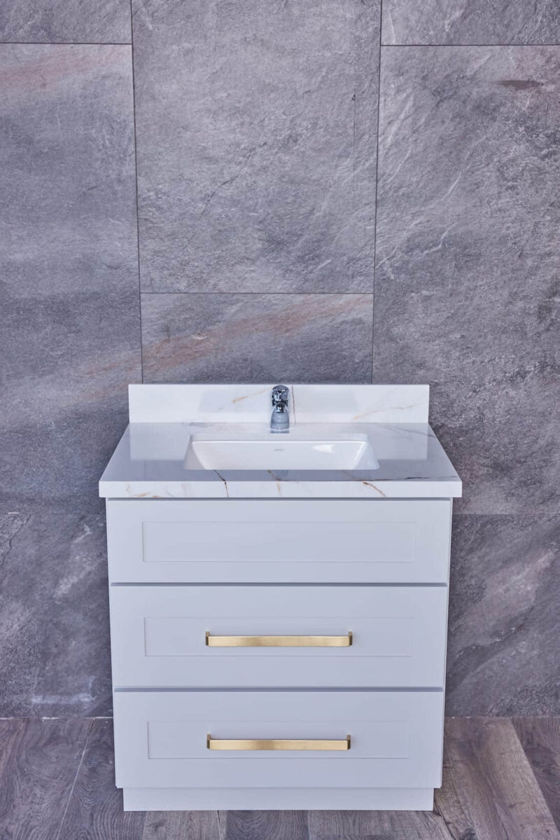 White bathroom vanity with marble countertop and gold handles against gray tile wall.