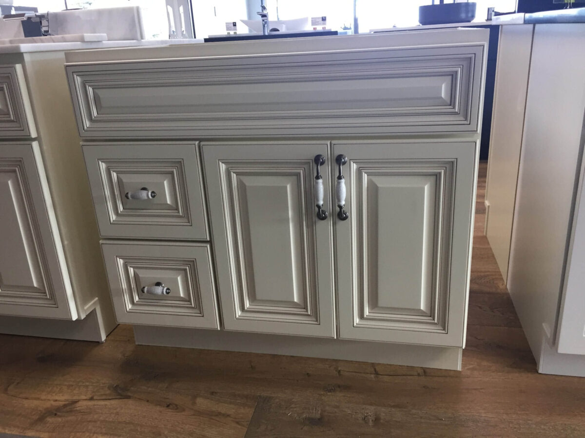 White kitchen cabinet with ornate paneling and silver handles on wooden floor.