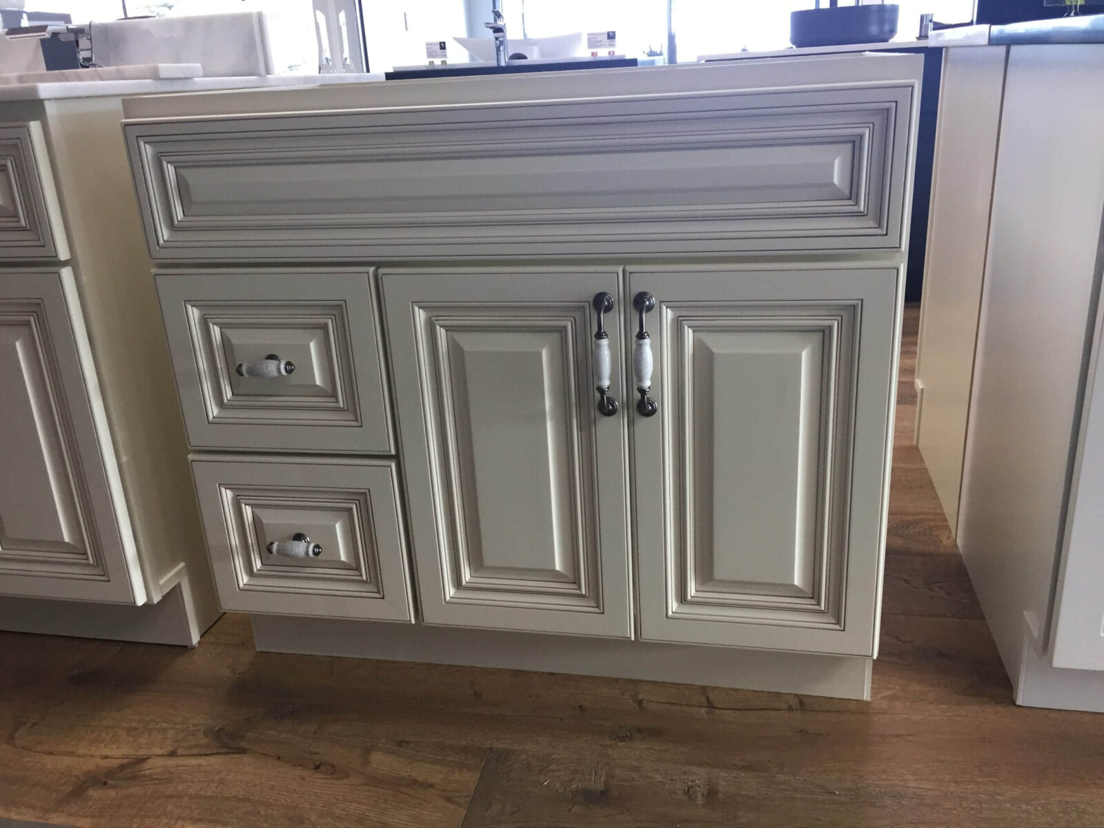 White kitchen cabinet with ornate paneling and silver handles on wooden floor.