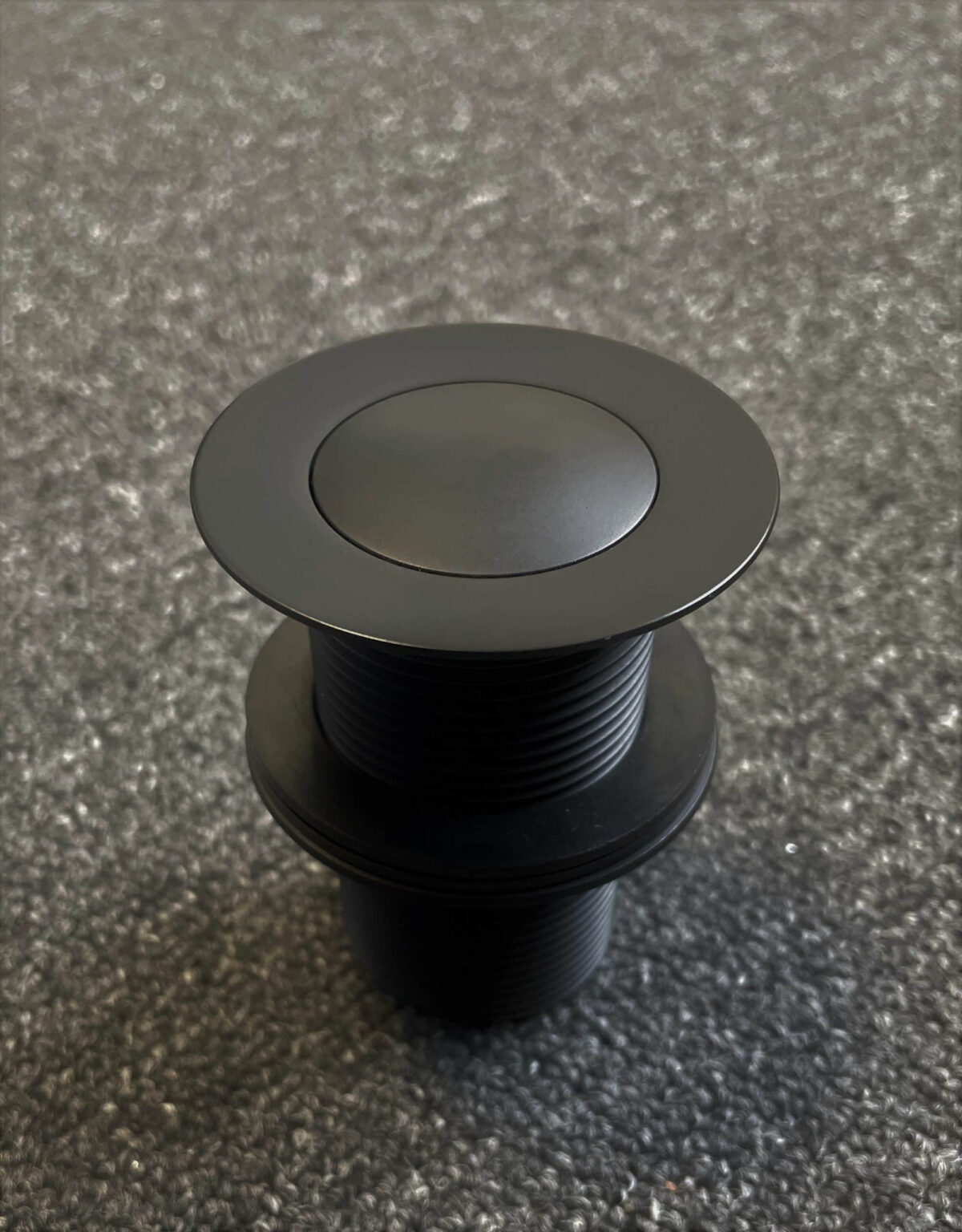 Matte black 32mm pop-up waste on textured surface.