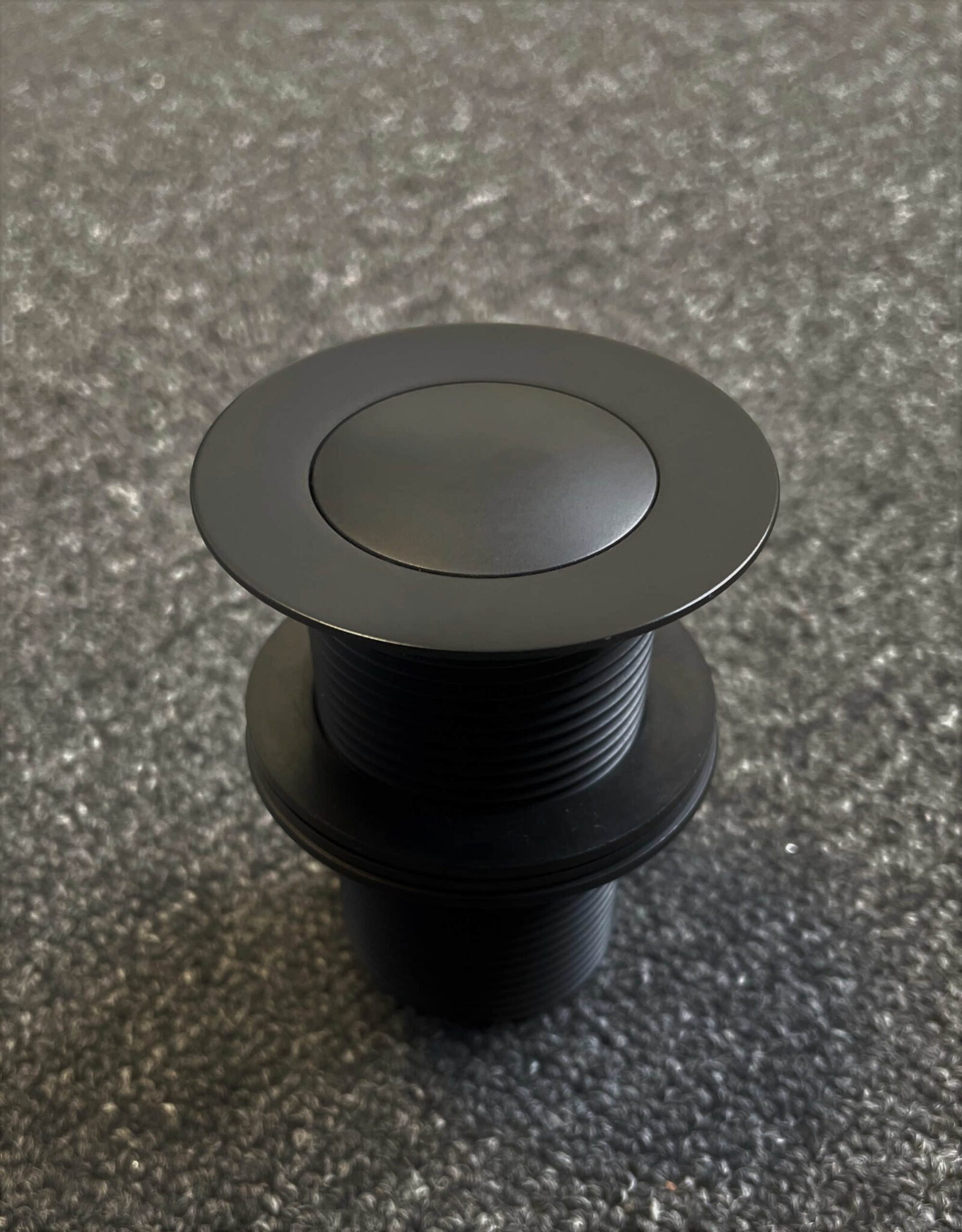 Matte black 32mm pop-up waste on textured surface.