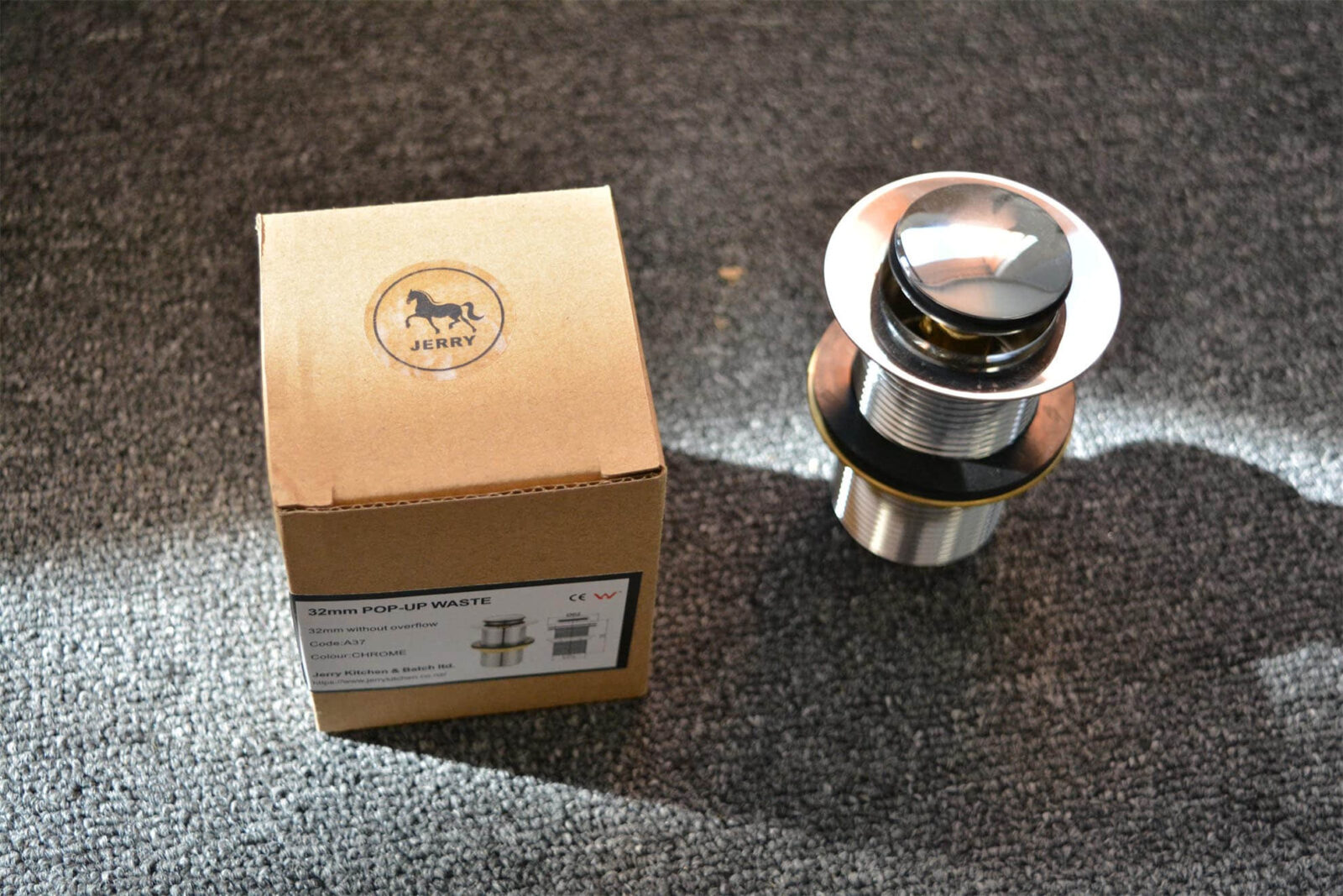 Chrome pop-up waste without overflow next to a cardboard box on carpet.