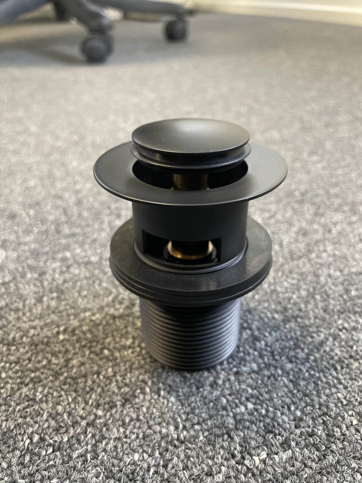 Matt black pop-up waste with overflow on carpeted floor.