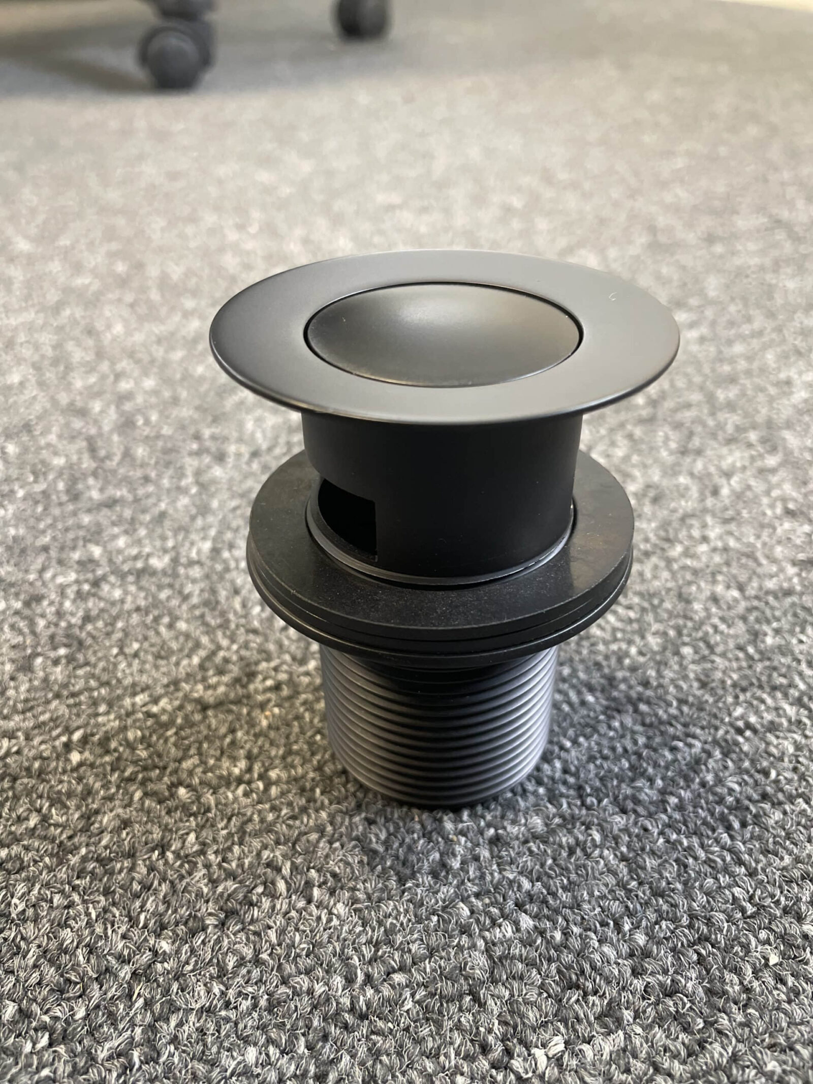 Matt black pop-up waste with overflow on gray surface.