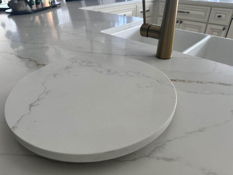 Marble kitchen countertop with round cutting board and gold faucet.