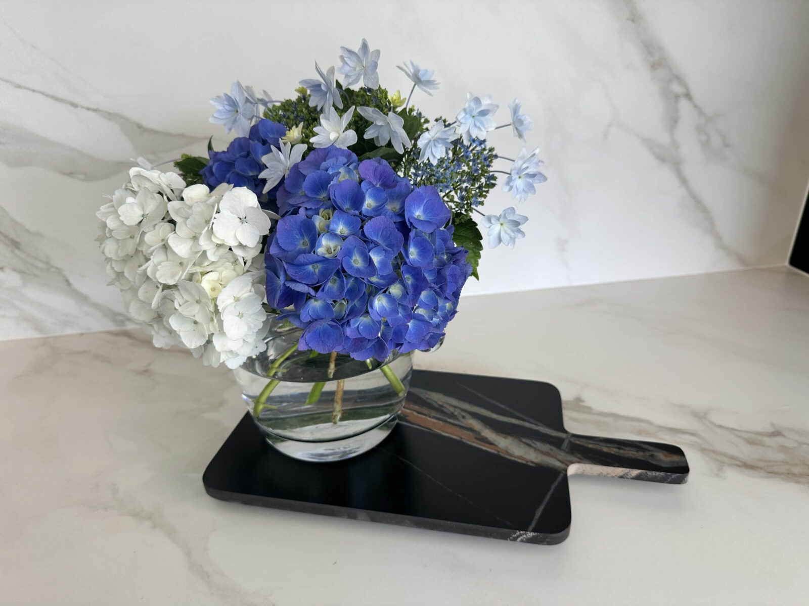 Blue and white hydrangeas in a glass vase on a marble countertop.