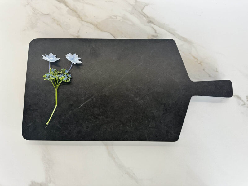 Black cutting board with two white flowers on a marble surface.