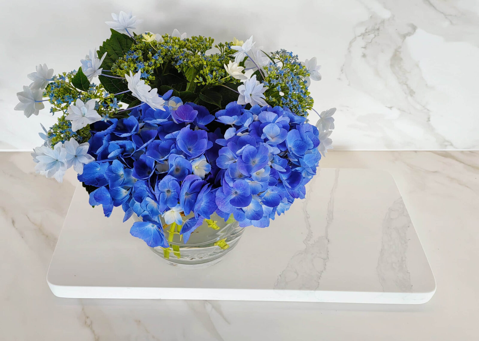 Blue hydrangeas in a glass vase on marble surface.