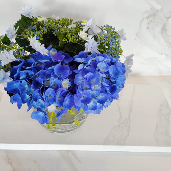Blue hydrangeas in a glass vase on marble surface.