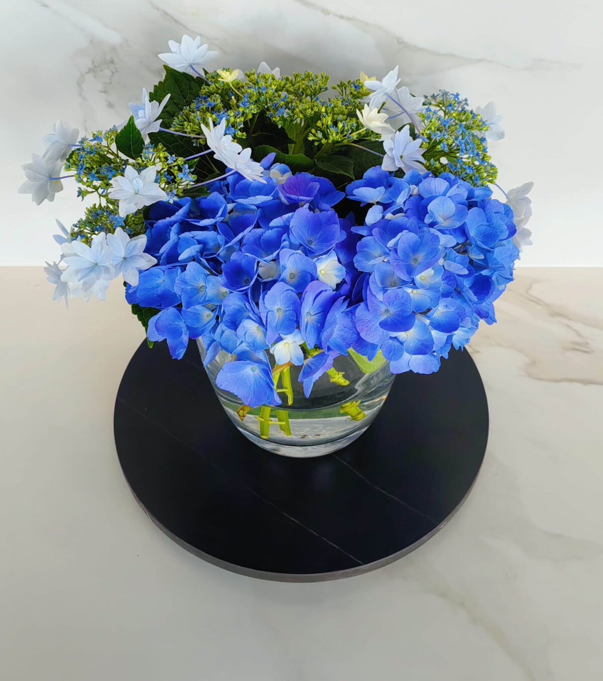 Blue hydrangea and white flowers in a glass vase on a black tray.