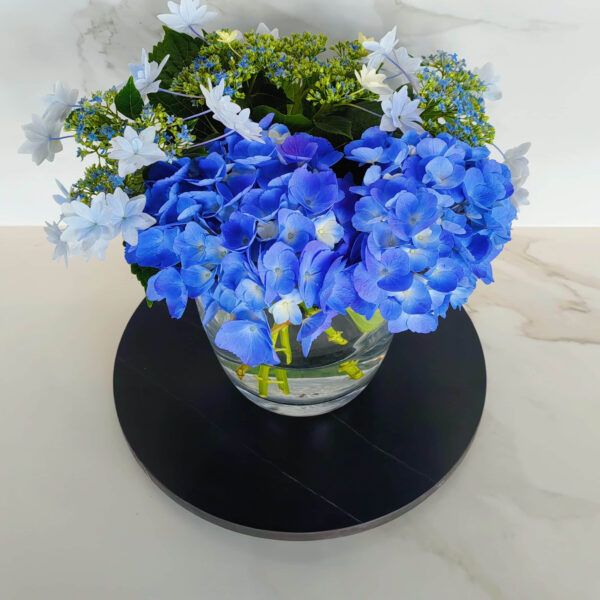 Blue hydrangea and white flowers in a glass vase on a black tray.