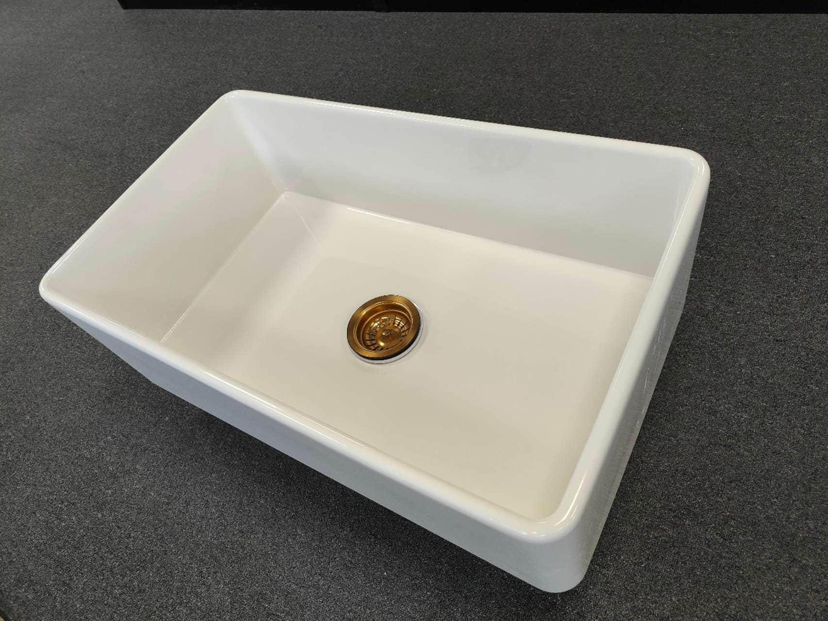 White rectangular kitchen sink with a gold drain on dark countertop.
